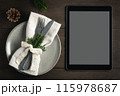 A black tablet with a blank screen on the table next to cutlery set out for Christmas dinner with a fork and knife. Mockup for design on the theme of Christmas holidays. 115978687