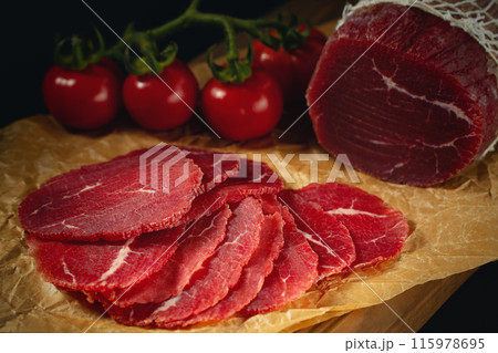 Whole and sliced bresaola on paper on a cutting board with tomatoes Whole and sliced bresaola on paper on a cutting board with tomatoes 115978695