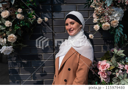 Portrait of a young caucasian woman dressed in a hijab near a wall with flowers. Portrait of a young caucasian woman dressed in a hijab near a wall with flowers. 115979480