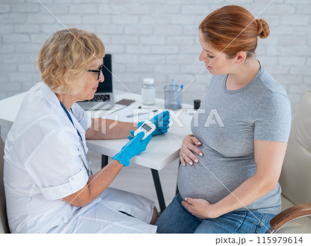 An elderly female doctor checks the blood sugar level of a pregnant red-haired woman. Gestational diabetes.  115979614