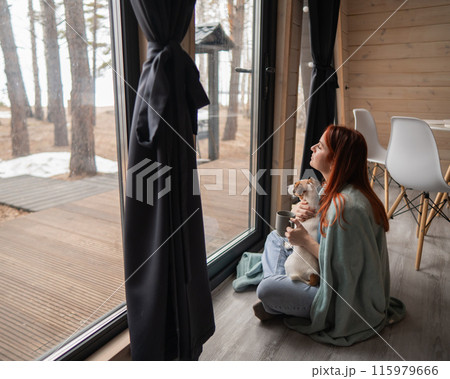 Caucasian woman wrapped in a blanket drinks coffee near the patio window hugging a Jack Russell terrier dog.  115979666