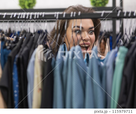 Portrait of a fat woman in a plus size store through hangers with clothes.  115979711