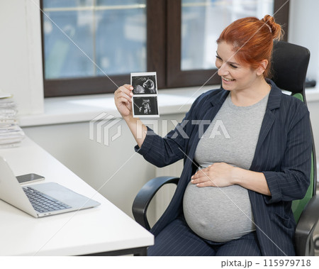 A pregnant woman shows a photo from an ultrasound scan of the fetus via video link in the office. 115979718