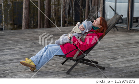 Caucasian woman with her son in an ergo backpack sitting in a wooden deck chair.  115979872