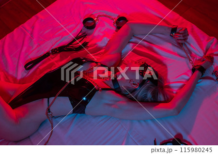 Sexy blonde woman in a leather mask and handcuffs and a collar lies in the bedroom in a red-blue neon light. Role-playing games. Sexy blonde woman in a leather mask and handcuffs and a collar lies in the bedroom in a red-blue neon light. Role-playing games. 115980245