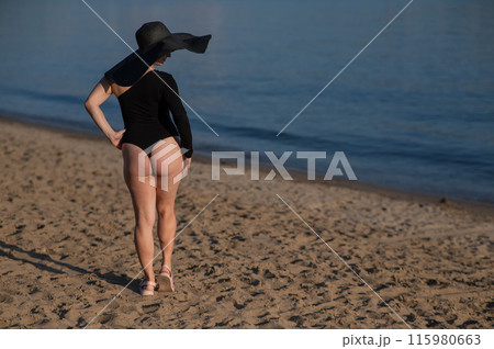 Rear view of woman in large straw hat and black swimsuit posing on the beach.  115980663
