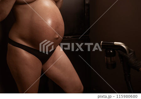 Close-up of the belly of a pregnant woman undergoing an instant tanning procedure. The master applies bronzer with a spray. 115980680