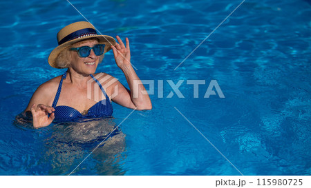 Portrait of an elderly woman in a hat and blue sunglasses swims in the pool.  115980725