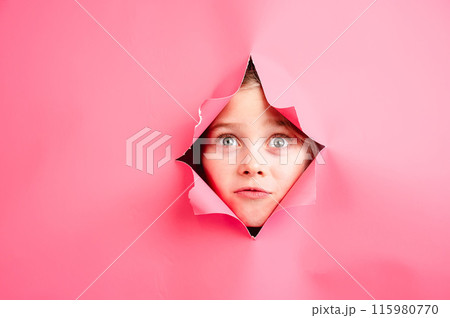 Cute Caucasian girl peeks out of a hole in a paper pink background. 115980770