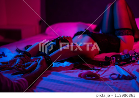 A woman in a leather mask and handcuffs lies on a bed in a blue-red light. Sex toys collection.  115980863