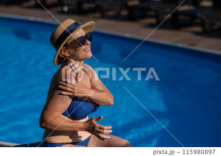 Portrait of an old woman in a straw hat, sunglasses and a swimsuit applying sunscreen to her skin while relaxing by the pool.  115980897