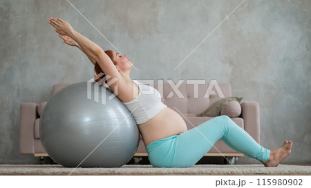 Pregnant red-haired woman doing exercises on fitness ball at home.  115980902