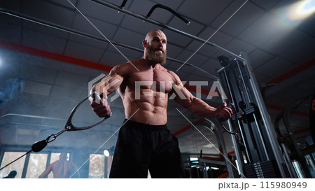 Bald Caucasian bodybuilder training chest using cable crossover in gym.  115980949