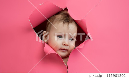 Cute Caucasian newborn baby boy peeks out of a hole in a paper pink background. 115981202