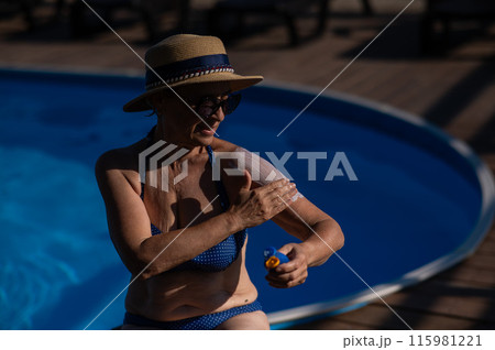 Portrait of an old woman in a straw hat, sunglasses and a swimsuit applying sunscreen to her skin while relaxing by the pool.  115981221
