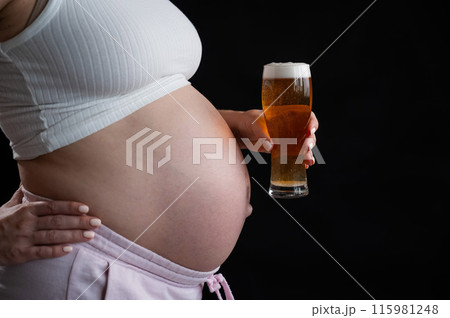 Close-up of the belly of a pregnant woman holding a glass of beer on a black background. Skin rash. Close-up of the belly of a pregnant woman holding a glass of beer on a black background. Skin rash. 115981248