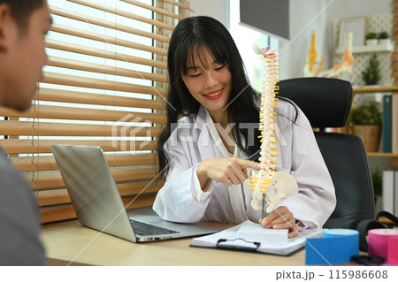 Smiling female Physical therapist showing anatomical model of spine, educating male patient in consultation room 115986608
