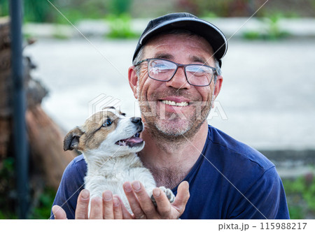 An old man who wears glasses with a skull on his head, holds in his hand a small and very beautiful dog, he plays with the dog and hugs it, it has a very beautiful smile. The dog is man's friend. An old man who wears glasses with a skull on his head, holds in his hand a small and very beautiful dog, he plays with the dog and hugs it, it has a very beautiful smile. The dog is man's friend. 115988217