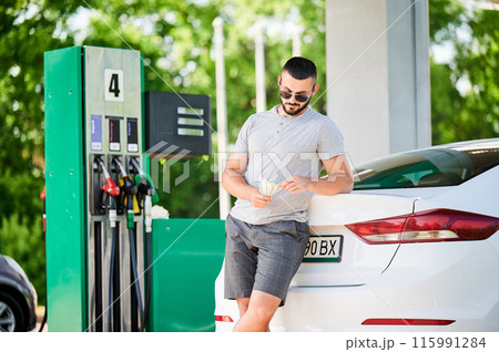 Young man waiting for petrol station operator to pay for fuel. Man leaning on his car and counting how much gasoline costs. Male in casual clothes holding money. Young man waiting for petrol station operator to pay for fuel. Man leaning on his car and counting how much gasoline costs. Male in casual clothes holding money. 115991284