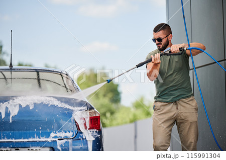 Man cleaning his car at self-service car wash with high pressure water jet. Confident man washing his blue auto with water gun at sunny day. 115991304