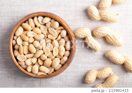 Roasted and salted peanuts in a wooden bowl on linen fabric. Ready-to-eat snack food, made from fruits of Arachis hypogaea, also known as groundnut, goober, pindar or monkey nut. Close-up from above. 115991415