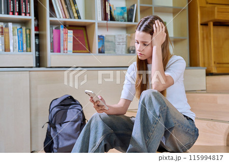 Sad teenage girl student looking at smartphone screen inside high school classroom 115994817
