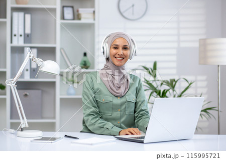 Smiling woman wearing hijab working on laptop at home office with headphones. Bright and modern workspace featuring white furniture, bookshelf, and plants. Concept of remote work and productivity Smiling woman wearing hijab working on laptop at home office with headphones. Bright and modern workspace featuring white furniture, bookshelf, and plants. Concept of remote work and productivity 115995721