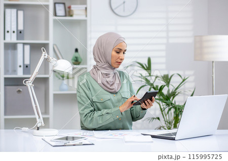 Female financial accountant calculates data with calculator while working at modern office desk with laptop and documents. Professional focusing on achieving accuracy in finances. 115995725