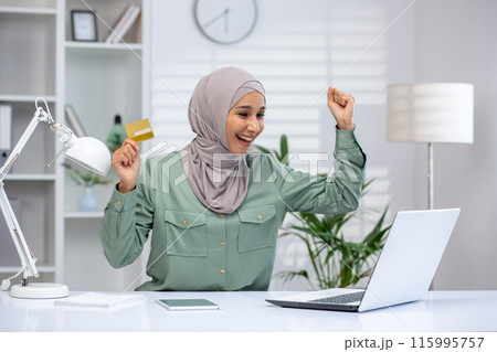 Woman in hijab excitedly raising arms while holding credit card, celebrating successful online transaction. Bright office setting with laptop, lamp, and shelves in background 115995757
