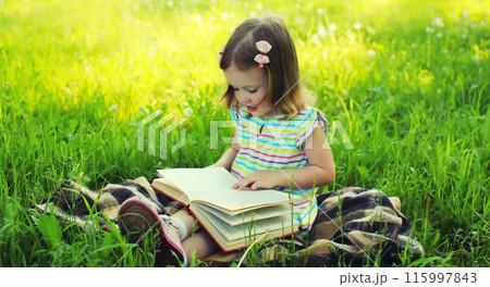 Happy little girl child reading book sitting on the grass in summer park 115997843