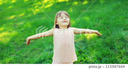Happy little girl child enjoying warm weather standing on the grass in summer green park 115997884