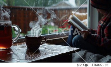 A rainy day scene: someone sits by the window, engrossed in a book, with a steaming cup of tea nearby. A rainy day scene: someone sits by the window, engrossed in a book, with a steaming cup of tea nearby. 116002256