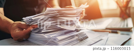 Person organizing a tall stack of papers on a desk in a bright office. Symbolizes work efficiency and document management. 116003015