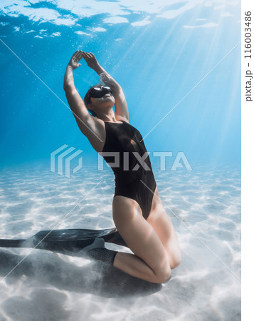 Woman underwater in blue ocean sitting on sandy sea bottom. Female posing with fins undersea 116003486