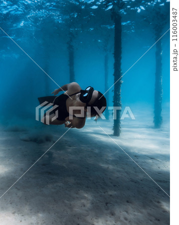 Freediver girl glides with fins dive under the pier in blue ocean. Female swims underwater dives near the pier pillars 116003487