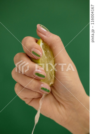 Vertical studio shot of womans hand with trendy nude nails decorated with green pattern squeezing fresh lime or lemon over green background, copy space 116004167