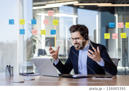 Businessman wearing suit and headset looking frustrated during video call in modern office. Sticky notes on glass wall. Concept of remote work, communication challenges, business frustration 116005391