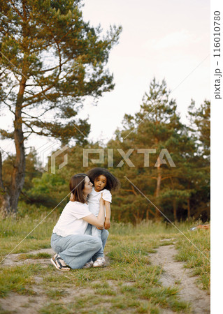 Photo of caucasian mother and her african american daughter embracing together outdoors. Girl has black curly hair. Mother and daugher wearing white t-shirts. Photo of caucasian mother and her african american daughter embracing together outdoors. Girl has black curly hair. Mother and daugher wearing white t-shirts. 116010780