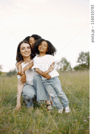 Photo of caucasian mother and two her african american daughters embracing together outdoors. Girl shas black curly hair. Mother and daughers wearing white t-shirts. Photo of caucasian mother and two her african american daughters embracing together outdoors. Girl shas black curly hair. Mother and daughers wearing white t-shirts. 116010785