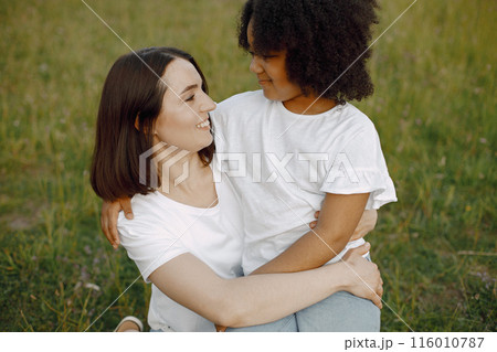 Photo of caucasian mother and her african american daughter embracing together outdoors. Girl has black curly hair. Mother and daugher wearing white t-shirts. 116010787