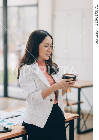 Beautiful young smiling Asian businesswoman working on laptop and drinking coffee, Asia businesswoman working document finance and calculator in her office. 116010971