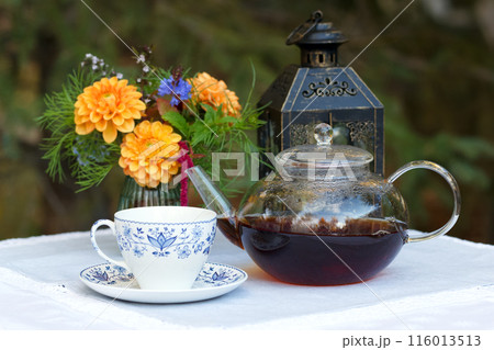 Patio table with cup of tea, pot, flowers and lantern. 116013513