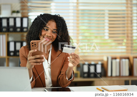Smiling African American female worker holding mobile phone and credit card for financial transaction Smiling African American female worker holding mobile phone and credit card for financial transaction 116016934