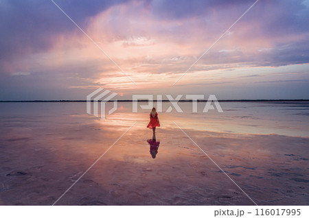 Aerial of a young woman in red dress walking in the water of a unique pink salt lake. Sunset at lake Bursol with beautiful reflections on calm water surface. Stunning scenery 116017995