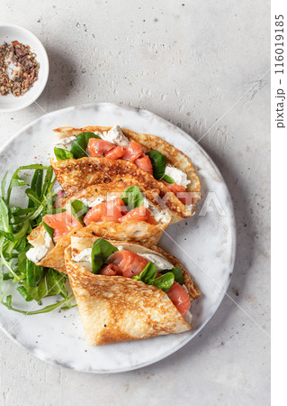 Crepes or thin pancakes with smoked salmon, soft cheese and spinach on a plate top view close up on gray textured background. Pancake day, Maslenitsa Crepes or thin pancakes with smoked salmon, soft cheese and spinach on a plate top view close up on gray textured background. Pancake day, Maslenitsa 116019185