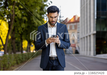 Businessman wearing suit using smartphone while walking in city street during autumn. Young professional checking messages and staying connected outdoors. Urban lifestyle ,modern technology concept 116025420