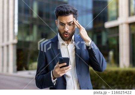 Shocked businessman in blue suit looking at smartphone with surprised expression outside modern office building, feeling shocked, stressed, and concerned while reading unexpected news or error. 116025422