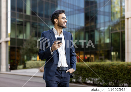Confident young businessman in suit holding smartphone and smiling while standing outdoors near modern office building. Professional attitude, business success, and positive outlook convey success 116025424
