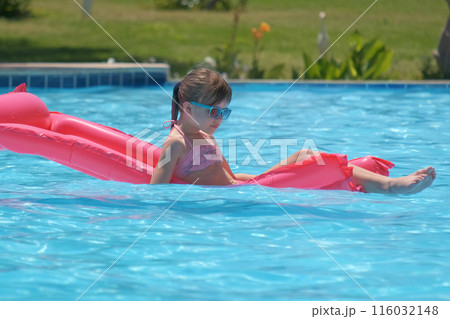 Happy child girl relaxing on inflatable air mattress in swimming pool on sunny summer day during tropical vacations. Summertime activities concept Happy child girl relaxing on inflatable air mattress in swimming pool on sunny summer day during tropical vacations. Summertime activities concept 116032148