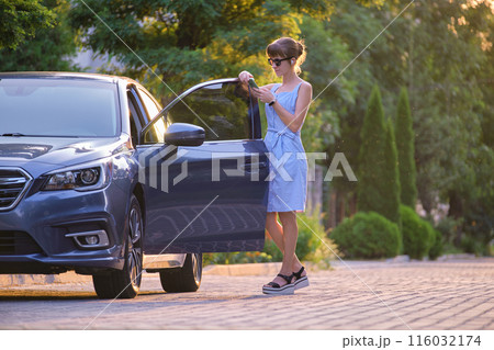 Young female driver standing near her car browsing her mobile phone on a city street in summer. 116032174